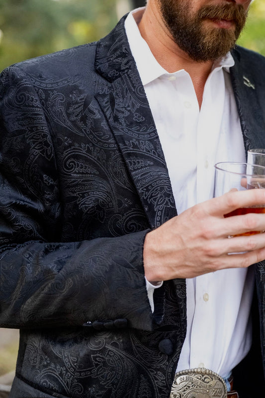 Man wearing a dark patterned blazer over a white shirt, holding a glass of amber liquid.