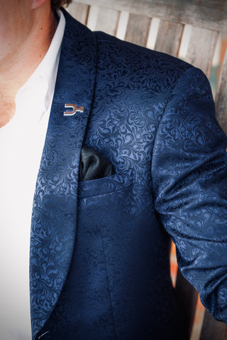 A close-up of a man wearing a textured navy blue patterned blazer over a white shirt, featuring a black pocket square and a small metallic lapel pin.