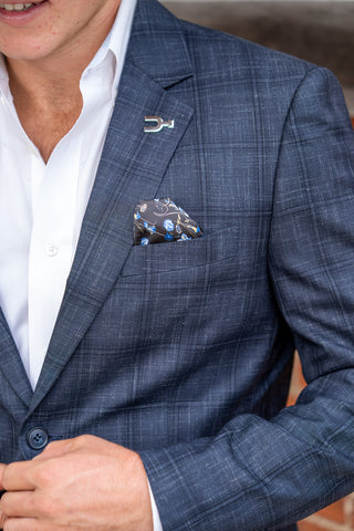 A close-up of a man wearing a blue plaid suit jacket over a white dress shirt, with a patterned pocket square in the breast pocket and a silver lapel pin.