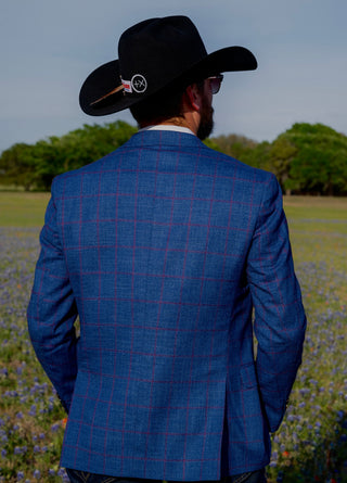 Man wearing a blue checkered suit and black cowboy hat in a field with trees in the background