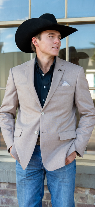A man wearing a light beige western blazer, black cowboy hat, dark button-down shirt, and blue jeans, standing in front of a window.