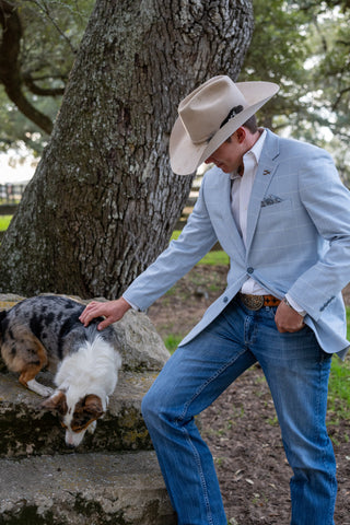 Man in light blue blazer and cowboy hat interacting with a dog by a tree