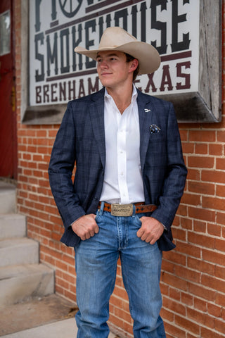 A man wearing a navy blue plaid blazer, white dress shirt, blue jeans, and a beige cowboy hat stands in front of a brick wall with a 'No Smokehouse' sign.