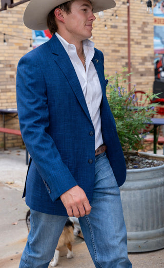 A man wearing a blue textured blazer, white dress shirt, blue jeans, and a cowboy hat walking outdoors in front of a brick wall and potted plants.