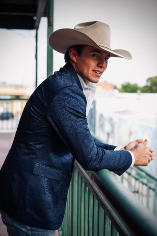A man wearing a beige cowboy hat, a textured navy blue blazer, and a white collared shirt leans against a green railing in an outdoor setting.