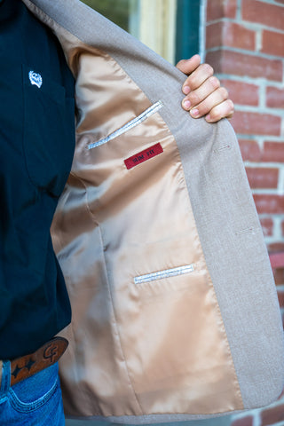 Close-up of a person holding open a light gray blazer to show the tan satin-lined interior, with a brick wall in the background.
