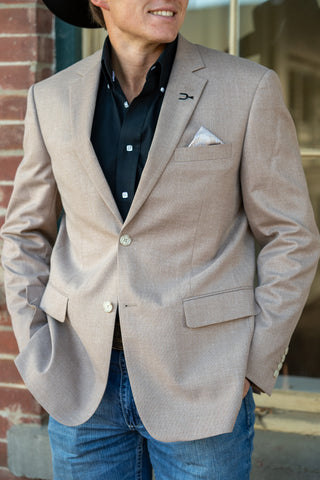 A man wearing a light beige textured blazer over a black button-down shirt and blue jeans, standing outdoors against a brick wall.