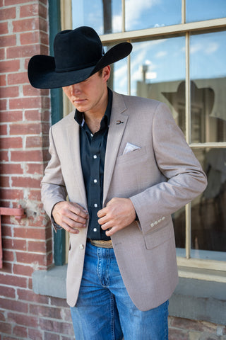 A man wearing a light beige blazer, black cowboy hat, dark button-down shirt, and blue jeans stands in front of a brick building window.