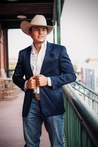 A man wearing a navy blue textured blazer, white dress shirt, blue jeans, and a beige cowboy hat, standing on an outdoor balcony.