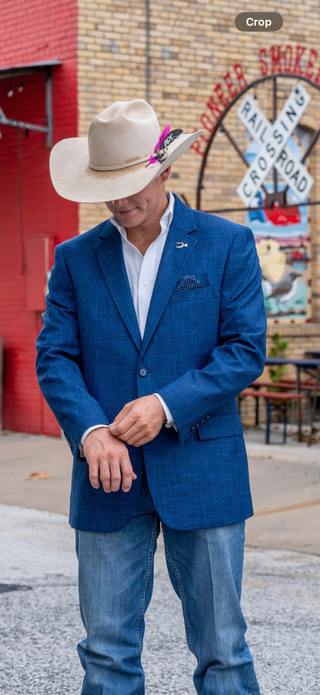 A man wearing a blue textured blazer, white dress shirt, blue jeans, and a beige cowboy hat with a purple feather, standing outdoors in front of a brick building with a railroad sign.