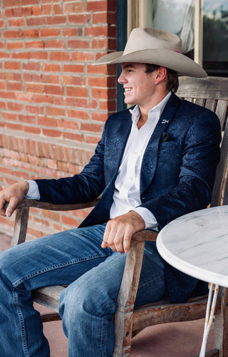 A man wearing a navy blue textured blazer, white dress shirt, blue jeans, and a beige cowboy hat sits smiling in a wooden rocking chair on a porch next to a brick wall.