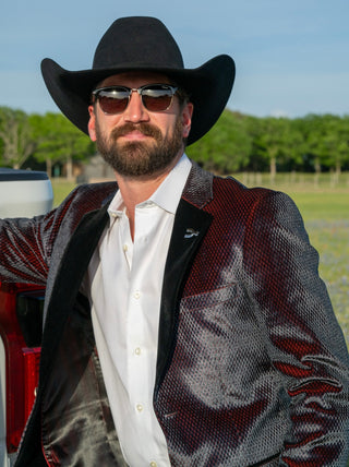 Man wearing a black cowboy hat and sunglasses, standing outdoors with trees in the background.