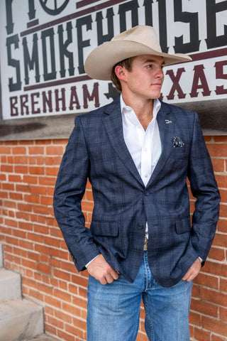 A man wearing a navy blue plaid blazer, white dress shirt, blue jeans, and a beige cowboy hat stands in front of a brick wall with a Western-themed sign that reads 'The Smokehouse Brenham Texas'.