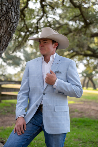 Man wearing a light blue blazer, white shirt, and cowboy hat in a natural setting with trees and grass.