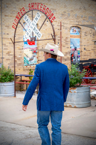 A man wearing a blue blazer, blue jeans, and a cream cowboy hat walks away from the camera in front of a brick building with a rustic 'Pioneer Smokehouse' sign.