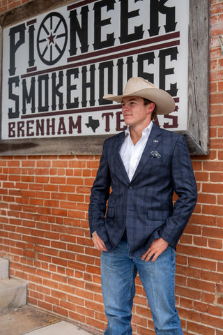 A man wearing a navy blue plaid blazer, white dress shirt, blue jeans, and a beige cowboy hat stands in front of a brick wall with a large 'Pioneer Smokehouse' sign.