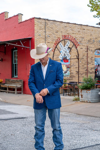A man wearing a blue blazer, jeans, and a beige cowboy hat stands on a street in front of a brick building with a red side building. The hat has a small purple flower accent.