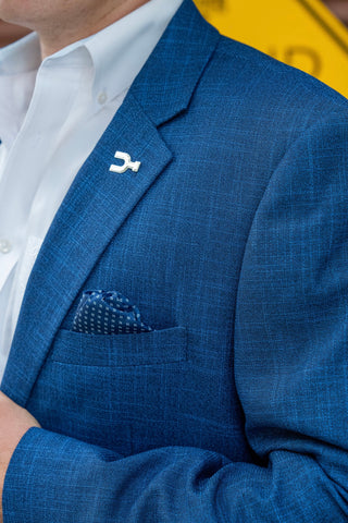 A close-up of a man wearing a textured blue suit jacket over a white dress shirt, featuring a small white lapel pin and a matching blue patterned pocket square.