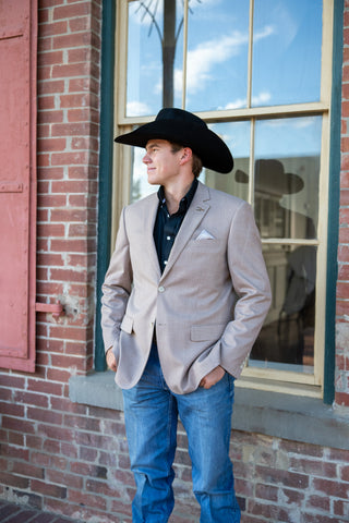 A man wearing a black cowboy hat, a light beige blazer, a dark button-down shirt, and blue jeans stands in front of a brick building window.