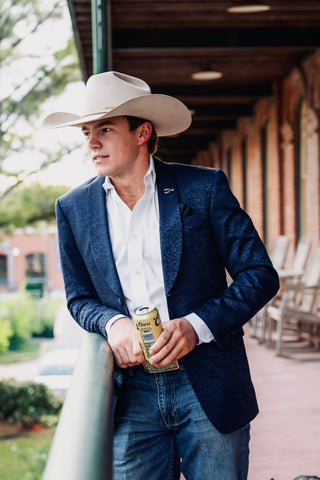 A man wearing a navy blue textured blazer, white dress shirt, blue jeans, and a cream cowboy hat stands on a porch holding a beer can.