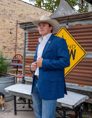 A man wearing a blue textured blazer, white dress shirt, and light blue jeans stands outdoors in front of a rustic corrugated metal structure with a yellow warning sign. He is also wearing a beige cowboy hat.