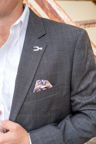 A close-up of a man wearing a dark gray micro-check suit jacket over a white dress shirt, featuring a silver-toned lapel pin and a patterned pocket square.