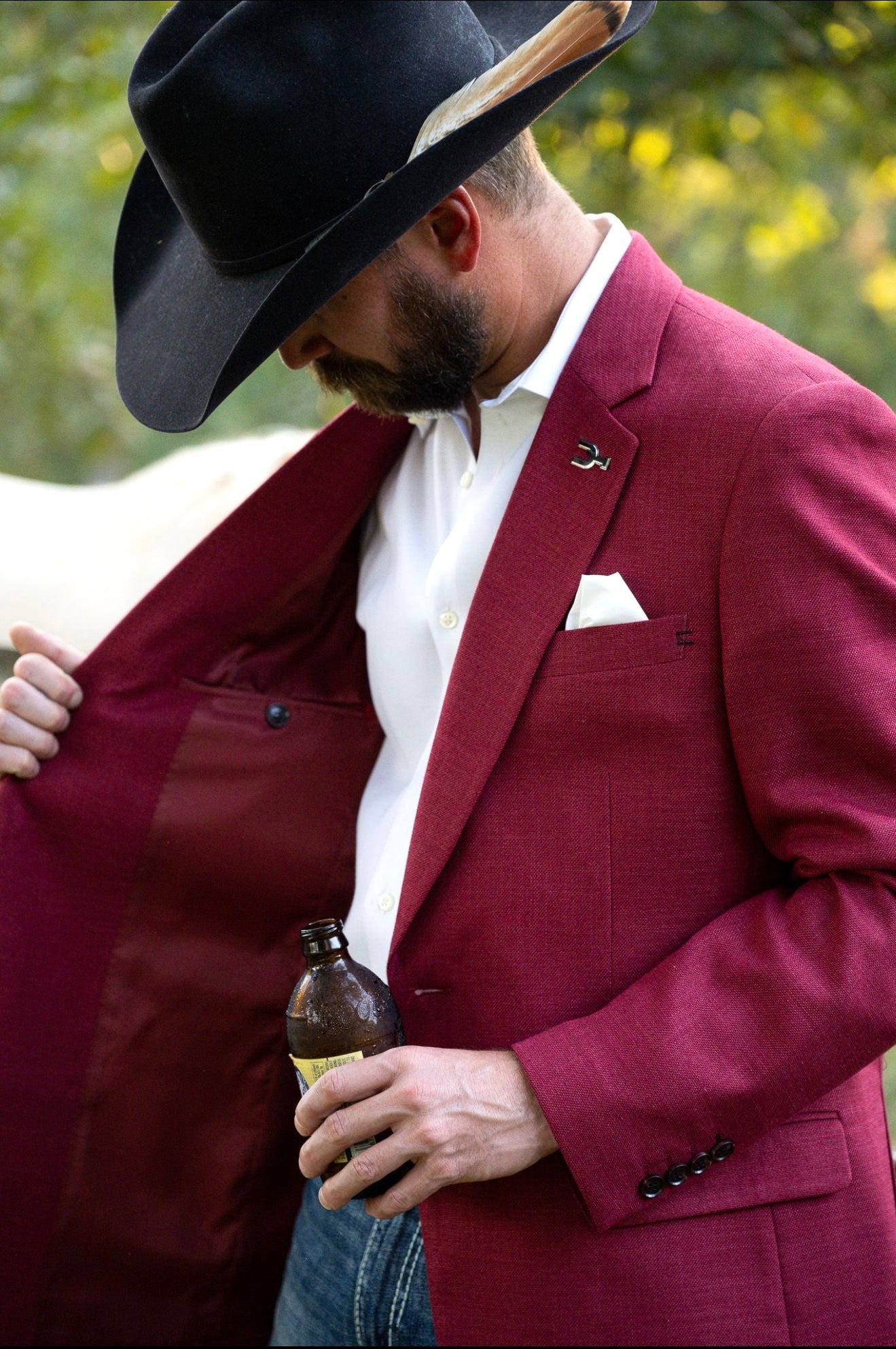 Man in a maroon suit and black cowboy hat holding a bottle, with a blurred natural background