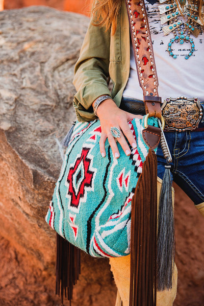 Saddle blanket purse, turquoise and held by a cowgirl wearing a belt buckle and chaps.